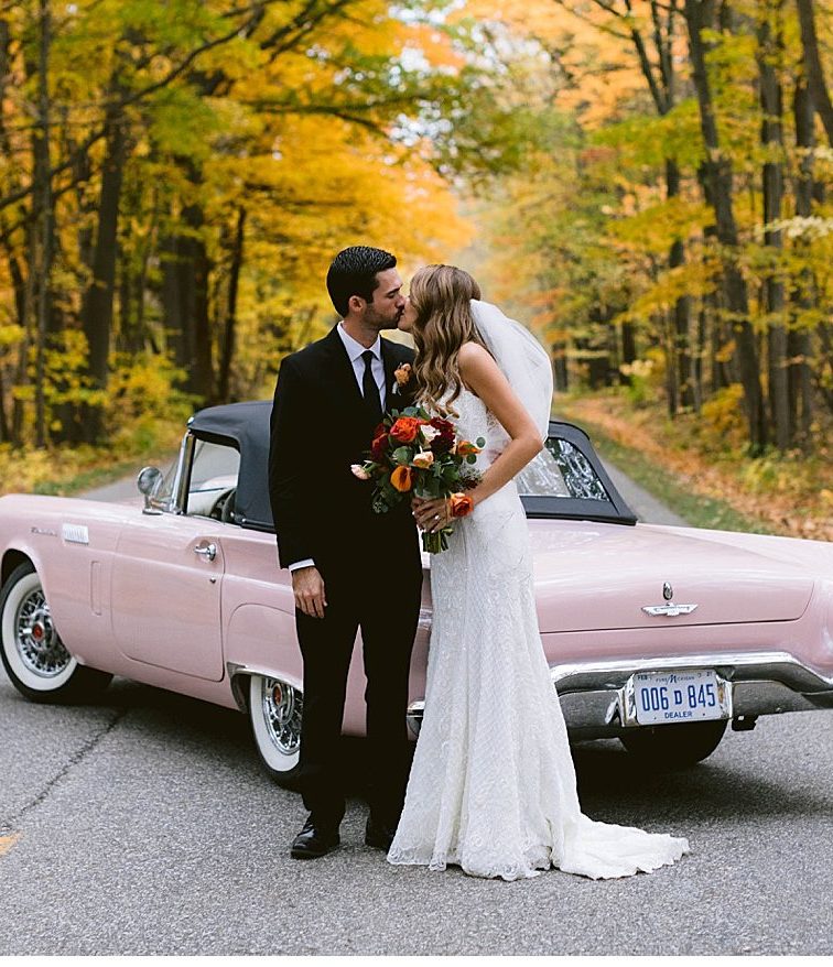 Couple kisses in the middle of a tunnel of fall trees with classic get away car behind them. Photo is very classic, elegant, and timeless taken in detroit michigan.
